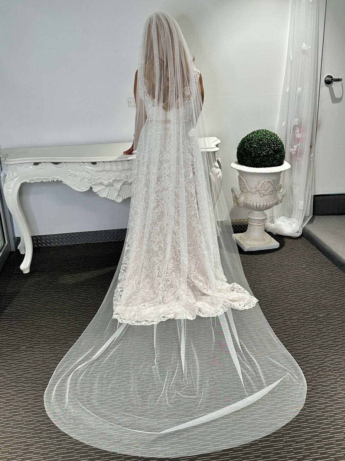 A woman standing in a room with a white bridal veil draped over her, reaching almost to the floor, with a lace trim at the bottom.