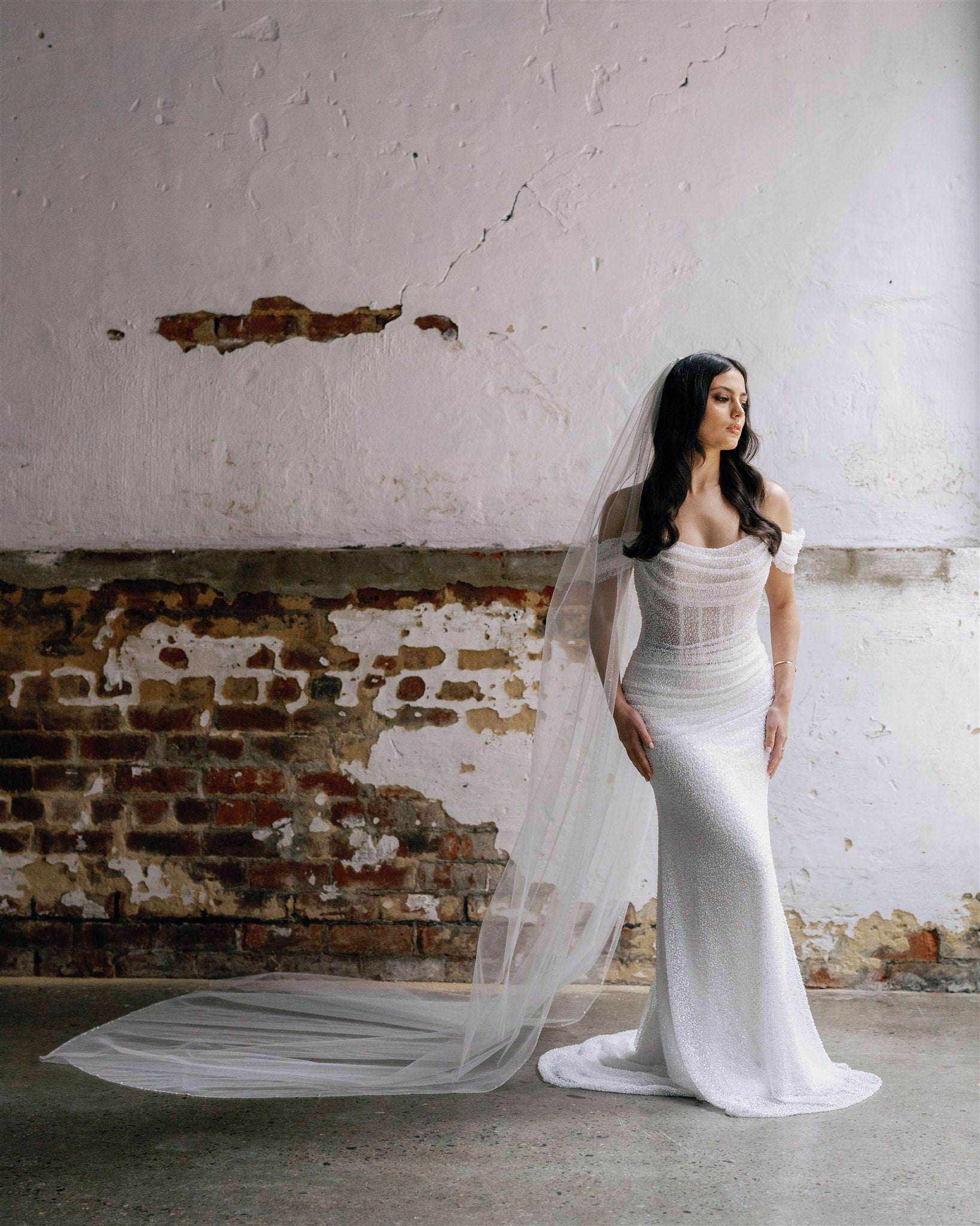 Bride stands in front of white painted brick wall wearing a beaded wedding dress and a cathedral length beaded veil 