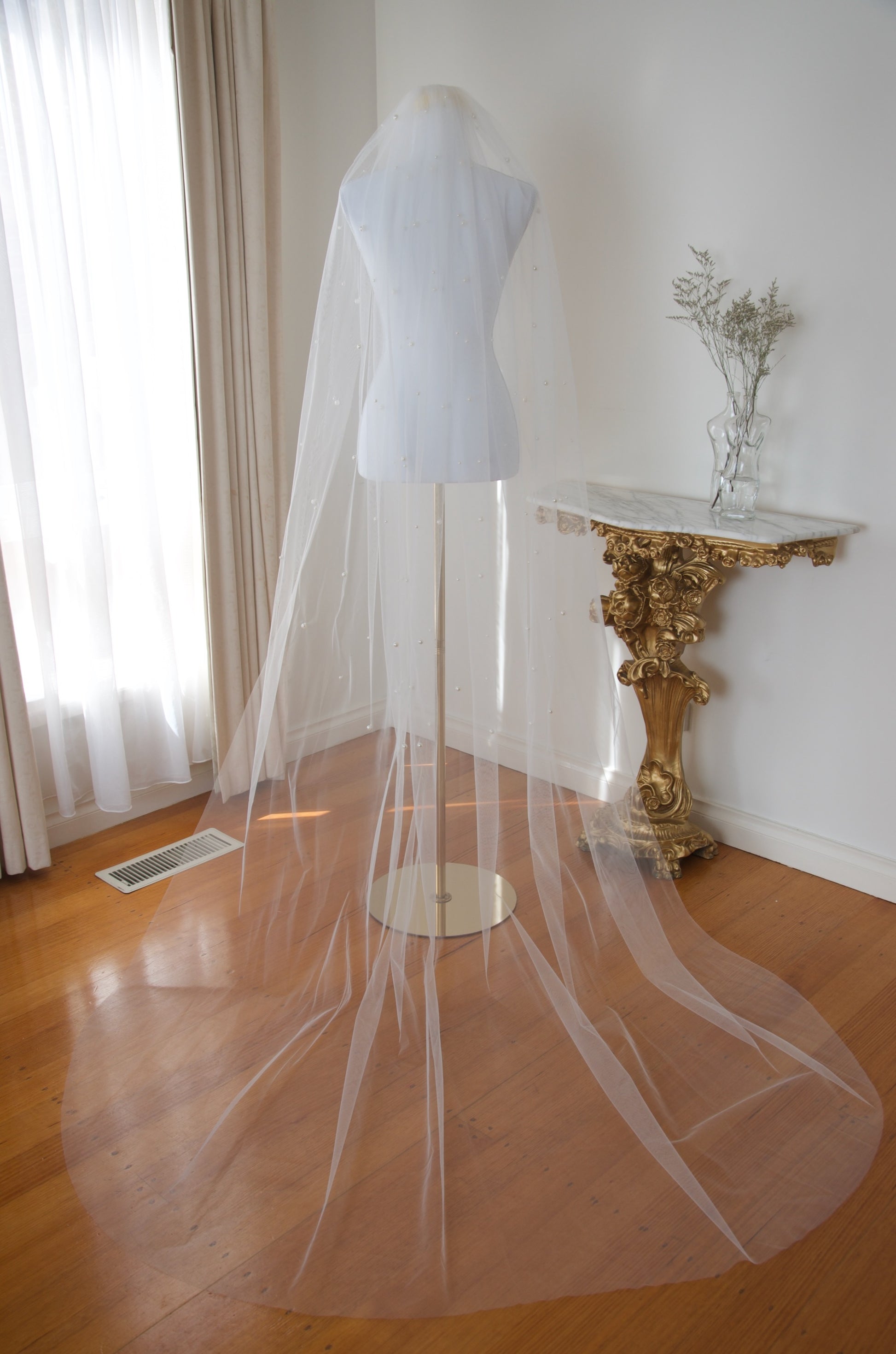 White wedding veil on a mannequin in a room with a gold console table and curtains.