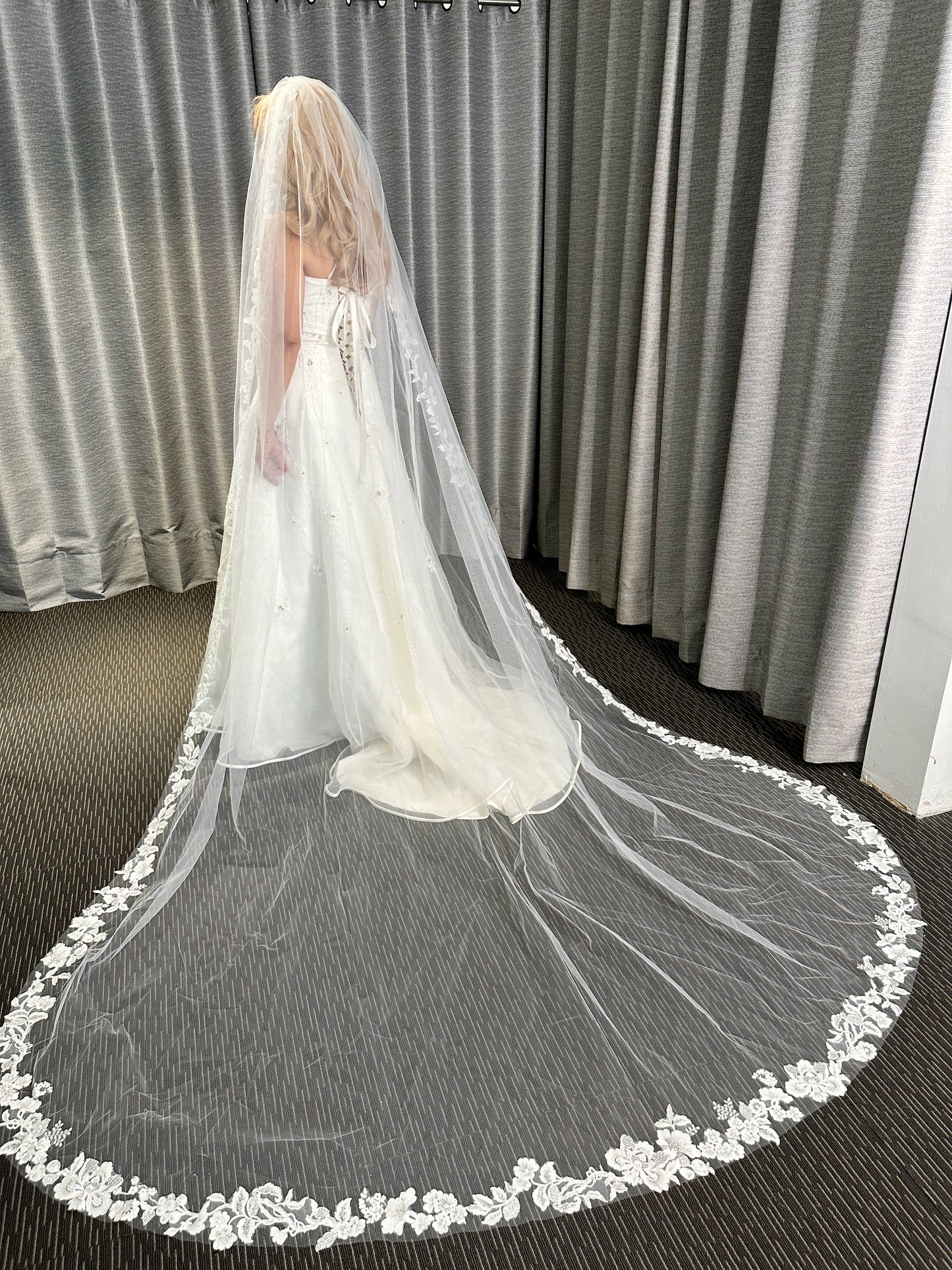 Bride wearing a long white wedding veil with floral lace details in front of gray curtains.