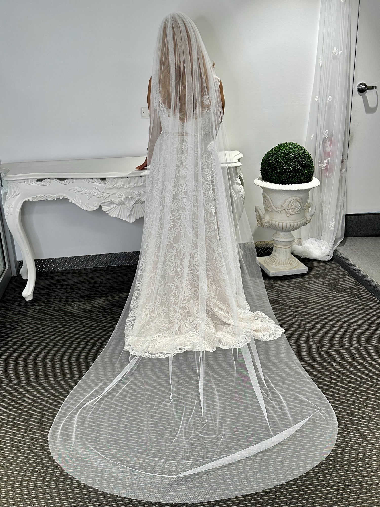 A woman standing in a room with a white bridal veil draped over her, reaching almost to the floor, with a lace trim at the bottom.