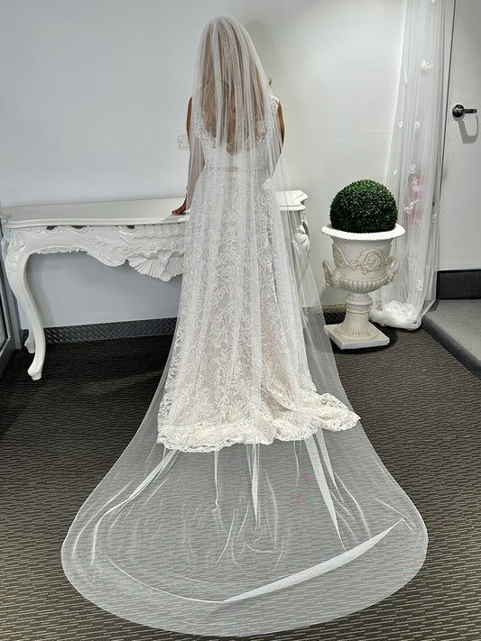 A woman standing in a room with a white bridal veil draped over her, reaching almost to the floor, with a lace trim at the bottom.